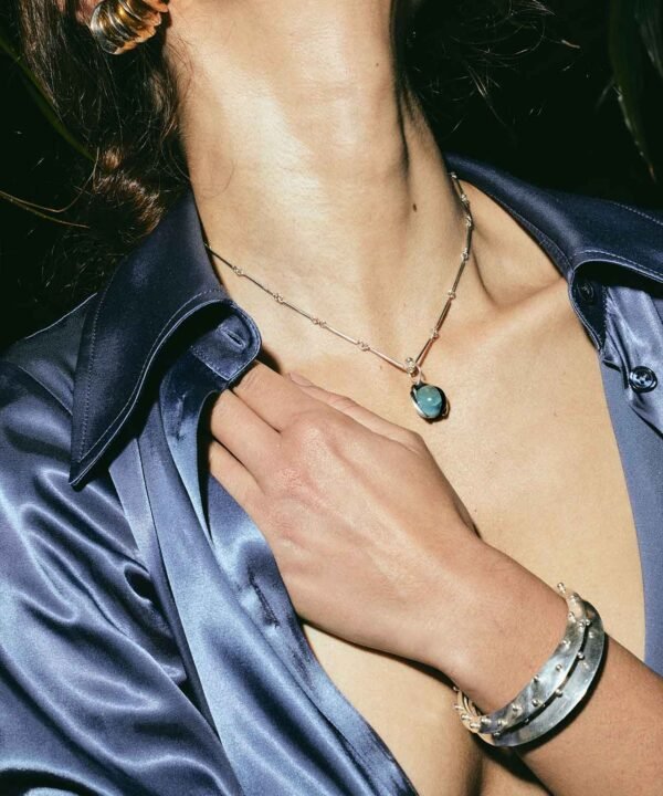 a woman caressing her shoulder with an open sating blue shirt. She is wearing a thisn link chains with round blue and black pendant and a stack of silver and gold ear cuffs. All jewelry is made by aysha bilgrami in recycled silver in bogota colombia