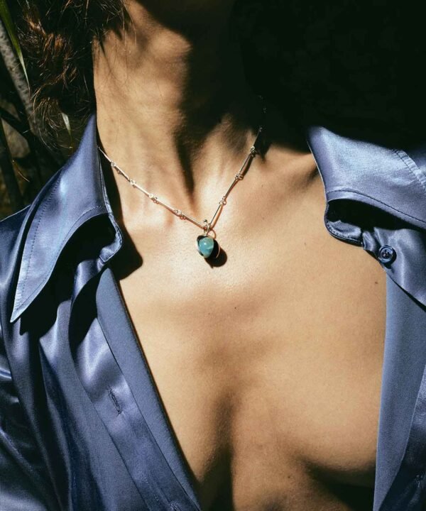 an uncovered womans chest wearing an open satin shirt and a thin chain and blue round pendant. Her jewelry is lady showing off 4 silver chains with point pendants dripping down her uncovered neck and 5 ear cuffs covering her ear. All her jewelry made by aysha bilgrami in recycled silver in bogota colombia
