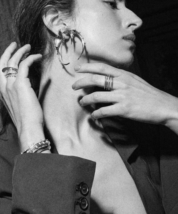 black and white side profile of woman reaching out to her neck whilst showing off three crescent shape hoops in three different sizes. She is also wearin a spiral silver ring in one hand and two rings in her other hand with two silver bangles. Her jewelry is made made by aysha bilgrami in recycled silver in bogota colombia
